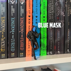 May include: A black figurine of a person wearing a blue mask and a hooded jacket, holding a book, stands in front of a row of books. The text "BLUE MASK" is displayed in white. The books have various titles and authors.