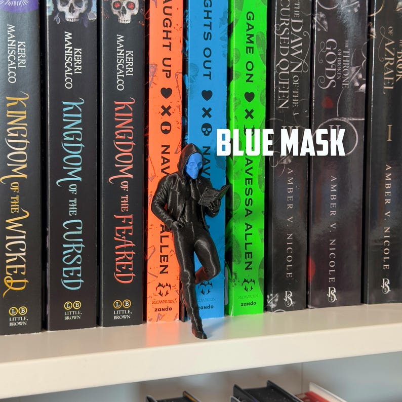 May include: A black figurine of a person wearing a blue mask and a hooded jacket, holding a book, stands in front of a row of books. The text "BLUE MASK" is displayed in white. The books have various titles and authors.