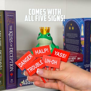 May include: A green frog figurine wearing a gold crown, holding five red signs with white text: "DANGER", "TROUBLE", "HALP!", "UH-OH", and "YASS!". The text "COMES WITH ALL FIVE SIGNS!" is above the frog.