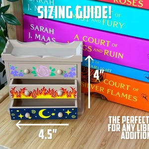 May include: A small, beige wooden drawer set with three drawers, each decorated with different designs. The top drawer has purple flowers and a pink rose, the middle drawer has flames, and the bottom drawer has a moon and stars. The set is 11.4 cm wide and 10.2 cm tall.