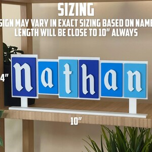 May include: A personalised name sign spelling "Nathan" in white, with each letter on a blue background. The sign is approximately 25 cm long and 10 cm tall. The text "SIZING" and other text are visible.