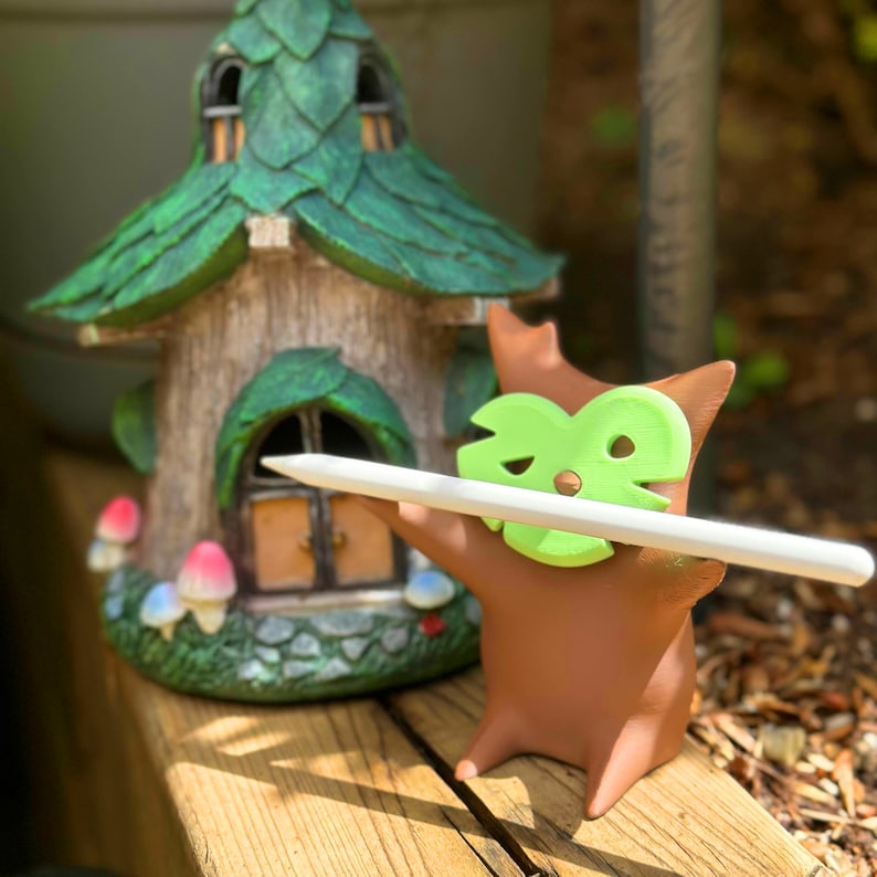 Puede incluir: Una figurita de cer&aacute;mica marr&oacute;n de una criatura con una m&aacute;scara verde en forma de hoja sosteniendo un l&aacute;piz blanco. La figurita est&aacute; de pie frente a una casa de hadas en miniatura verde y marr&oacute;n.