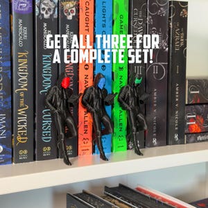 May include: Three black figurines with colored masks (red, blue, and green) are displayed in front of a row of books. The text "GET ALL THREE FOR A COMPLETE SET!" is visible. The books have titles like "Kingdom of the Wicked".