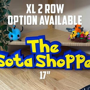 May include: A blue and yellow sign reading "The Sota Shoppe" is 43 cm long and 15 cm tall. The text "XL 2 ROW OPTION AVAILABLE" is above the sign. Small toys are on the shelf behind the sign.