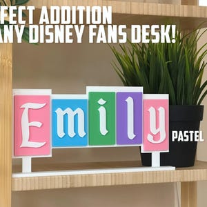 May include: A pastel-coloured nameplate with the name "Emily" in a white, stylised font. The nameplate is displayed on a white base, with a potted plant in the background. The text "PERFECT ADDITION TO ANY DISNEY FANS DESK!" is at the top.