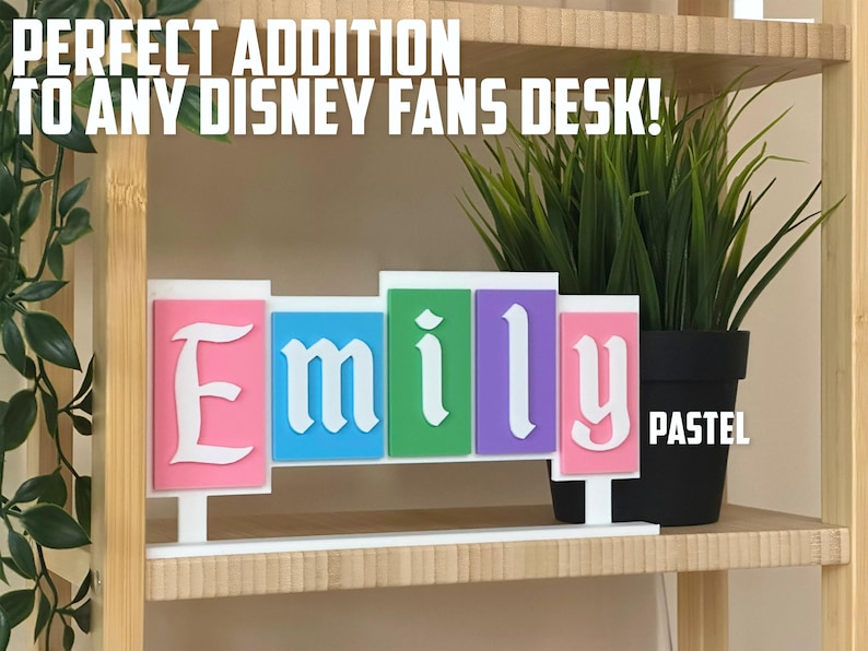 May include: A pastel-coloured nameplate with the name "Emily" in a white, stylised font. The nameplate is displayed on a white base, with a potted plant in the background. The text "PERFECT ADDITION TO ANY DISNEY FANS DESK!" is at the top.