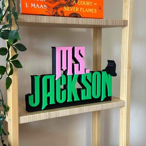 May include: A wooden shelf with a pink and green sign that reads "Ms. Jackson". The sign is made of 3D letters and is sitting on a shelf with a book titled "A Court of Silver Flames" by J. Maas.