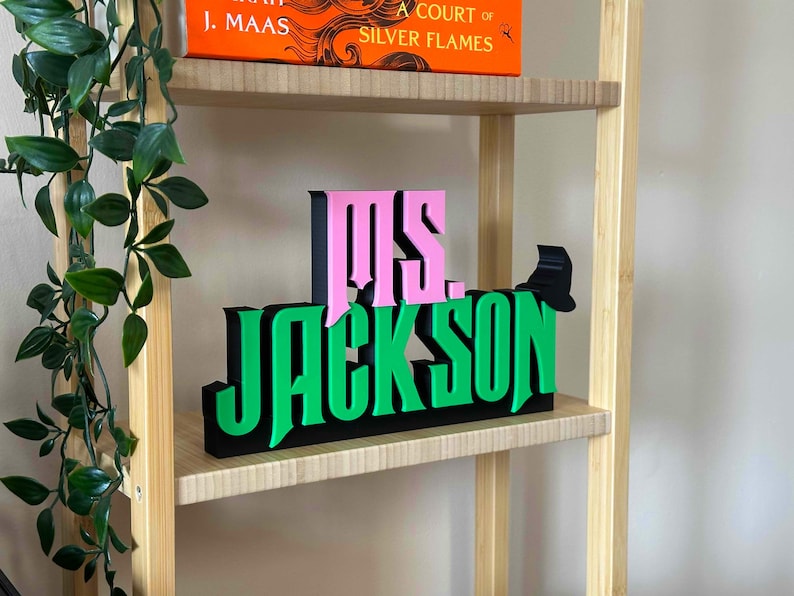 May include: A wooden shelf with a pink and green sign that reads "Ms. Jackson". The sign is made of 3D letters and is sitting on a shelf with a book titled "A Court of Silver Flames" by J. Maas.