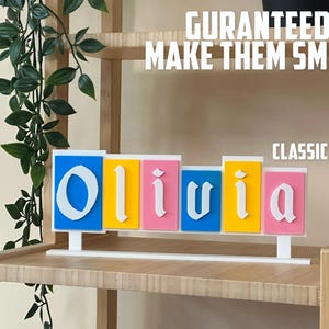 May include: A decorative name sign spelling "Olivia" in white, block letters. The letters are on separate rectangular blocks in blue, yellow, and pink. The sign is on a white base, with the text "GURANTEED TO MAKE THEM SMILE!" above.