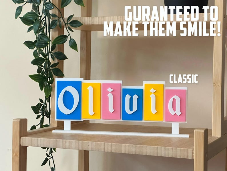 May include: A decorative name sign spelling "Olivia" in white, block letters. The letters are on separate rectangular blocks in blue, yellow, and pink. The sign is on a white base, with the text "GURANTEED TO MAKE THEM SMILE!" above.