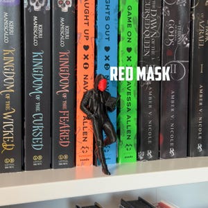May include: A black figurine of a person wearing a red mask and a hooded jacket, leaning against a bookshelf filled with colorful books. The text "RED MASK" is displayed in white.