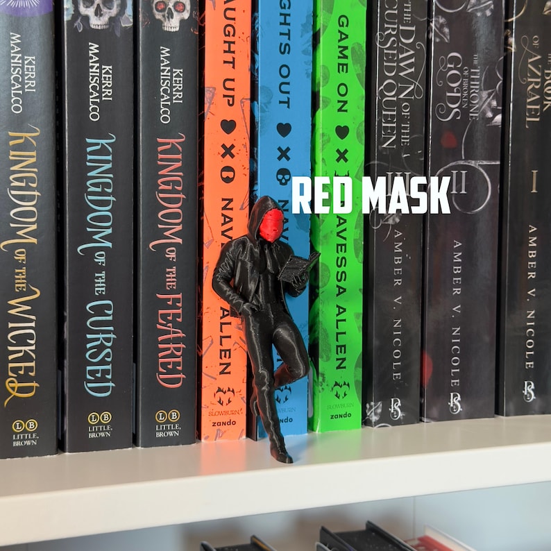 May include: A black figurine of a person wearing a red mask and a hooded jacket, leaning against a bookshelf filled with colorful books. The text "RED MASK" is displayed in white.