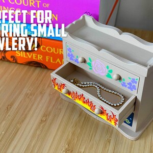 May include: A small, light-coloured wooden jewellery chest with two drawers. The top drawer is decorated with floral designs and the bottom drawer has a flame design. A necklace is inside the open drawer. The text "PERFECT FOR STORING SMALL JEWLERY!" is visible.