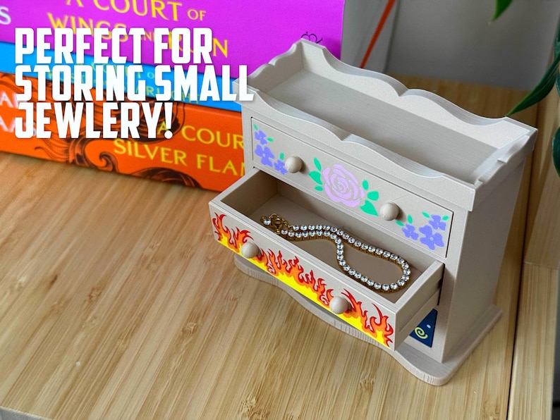 May include: A small, light-coloured wooden jewellery chest with two drawers. The top drawer is decorated with floral designs and the bottom drawer has a flame design. A necklace is inside the open drawer. The text "PERFECT FOR STORING SMALL JEWLERY!" is visible.