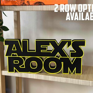 Peut inclure: Une enseigne en bois noire et jaune avec l'inscription "ALEX'S ROOM" sur une &eacute;tag&egrave;re en bois. L'enseigne est en deux rang&eacute;es avec "ALEX'S" sur la rang&eacute;e du haut et "ROOM" sur la rang&eacute;e du bas. Le texte "2 ROW OPTION AVAILABLE!" est &eacute;crit en blanc sur fond noir au-dessus de l'enseigne.