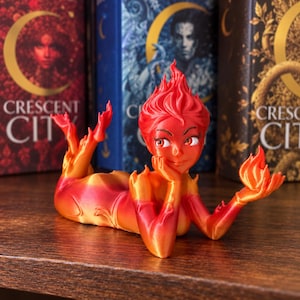 May include: A figurine of a humanoid character made of swirling red and orange flame-like material, lying in a reclining pose with one hand supporting the head and the other holding a small flame, featuring stylised hair and facial features.