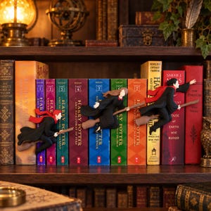 Könnte beinhalten: Drei Miniaturfiguren von Charakteren auf Besenstielen sind an den Buchrücken einer Reihe von Harry-Potter-Büchern befestigt. Die Figuren tragen schwarze Umhänge mit roten Schals und sind so positioniert, als würden sie horizontal fliegen.