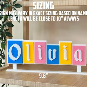 May include: A colourful name sign spelling "Olivia" in white, with each letter on a different coloured background: blue, yellow, pink, blue, and pink. The sign is 25 cm long and 10 cm tall.