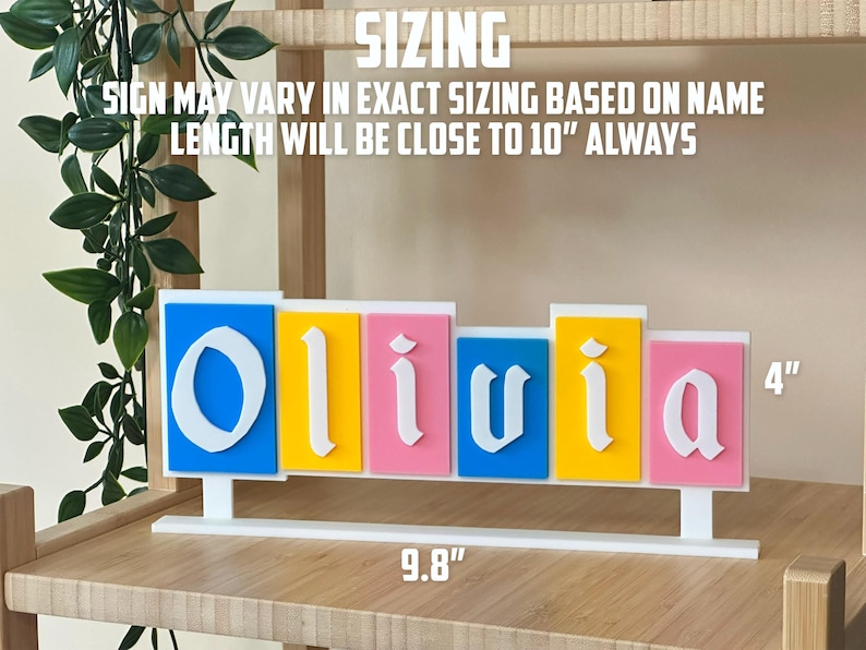 May include: A colourful name sign spelling "Olivia" in white, with each letter on a different coloured background: blue, yellow, pink, blue, and pink. The sign is 25 cm long and 10 cm tall.