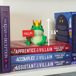 May include: A green frog figurine wearing a gold crown and holding a red sign that says "HALP!" The frog is 3.6 inches tall and sits atop a stack of books titled "Apprentice to the Villain", "Accomplice to the Villain", and "Assistant to the Villain".