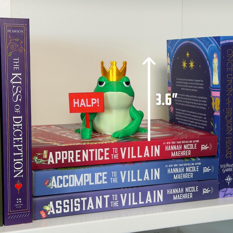 May include: A green frog figurine wearing a gold crown and holding a red sign that says "HALP!" The frog is 3.6 inches tall and sits atop a stack of books titled "Apprentice to the Villain", "Accomplice to the Villain", and "Assistant to the Villain".