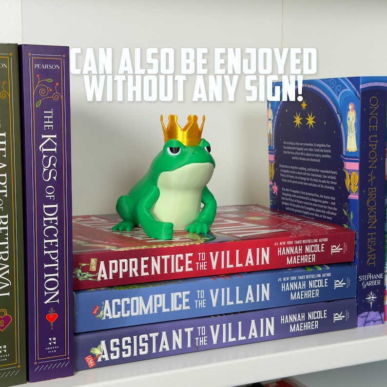 May include: A green frog figurine wearing a gold crown sits atop a stack of books. The books are titled "Apprentice to the Villain", "Accomplice to the Villain", and "Assistant to the Villain". The text "Can also be enjoyed without any sign!" is visible.
