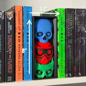 May include: A shelf displays a black box containing three stacked, colorful skull-shaped sculptures. The top skull is blue, the middle is red, and the bottom is green. The box is 3.2 inches wide and 8 inches tall. Books with titles like "Kingdom of the Feared" and "Lights Out" flank the box.