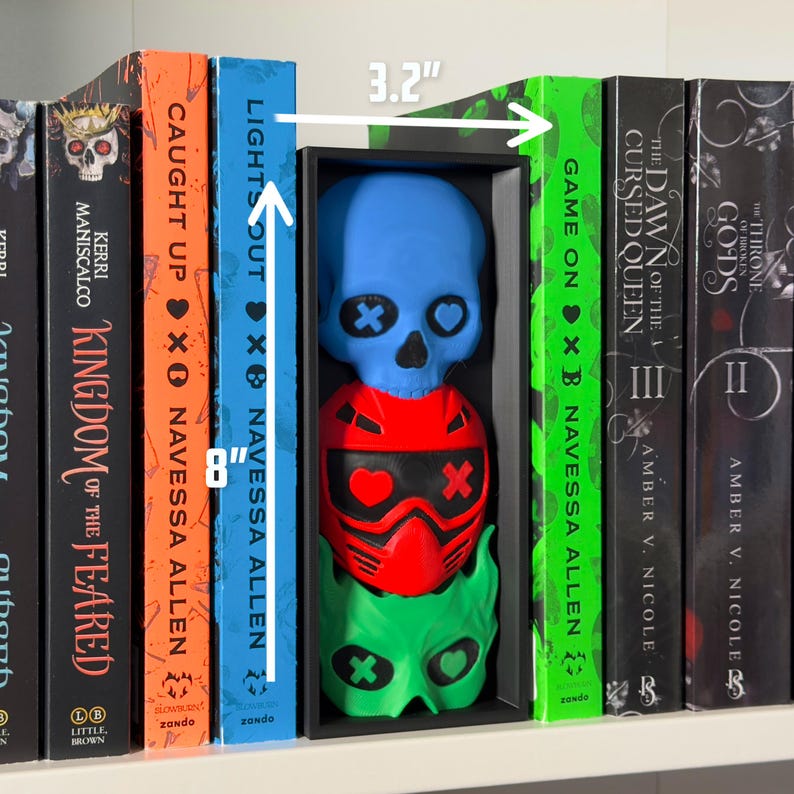 May include: A shelf displays a black box containing three stacked, colorful skull-shaped sculptures. The top skull is blue, the middle is red, and the bottom is green. The box is 3.2 inches wide and 8 inches tall. Books with titles like "Kingdom of the Feared" and "Lights Out" flank the box.