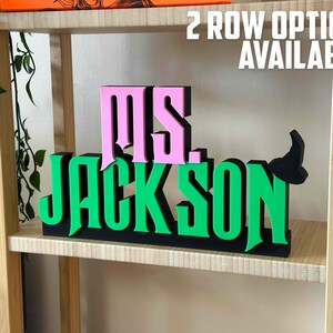 May include: A wooden sign with the words "MS. JACKSON" in pink and green letters. The sign is on a shelf with the text "2 ROW OPTIONS AVAILABLE!" above it.