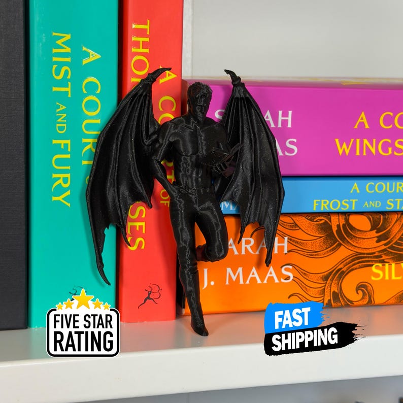 May include: A black figurine of a winged male figure, possibly a demon or angel, stands in front of a shelf of colorful books. The figurine has outstretched wings and is posed in a dynamic stance. The books have titles like "A Court of Mist and Fury".