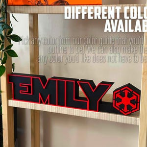 Peut inclure: Une &eacute;tag&egrave;re en bois avec une enseigne de nom 3D noire et rouge qui dit "EMILY" avec un symbole rouge et noir &agrave; c&ocirc;t&eacute;. Le texte "DIFFERENTES COULEURS DISPONIBLES !" est en haut de l'image. Le texte sous l'enseigne dit "Choisissez n'importe quelle couleur dans notre guide des couleurs que vous souhaitez pour le contour ! Nous pouvons &eacute;galement faire le nom dans n'importe quelle couleur que vous souhaitez, il ne doit pas n&eacute;cessairement &ecirc;tre noir !"