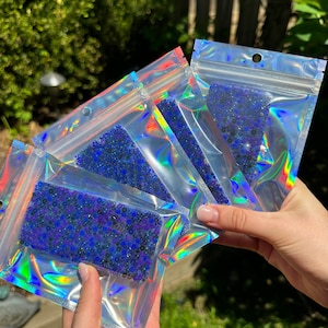 May include: Four iridescent clear plastic bags filled with blue and purple iridescent beads. The bags are stacked on top of each other.
