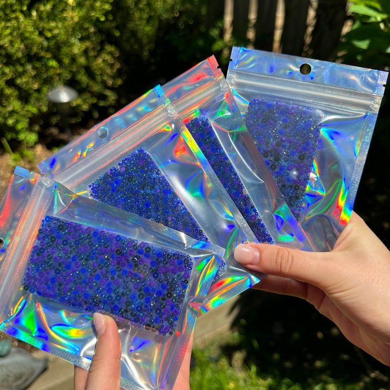 May include: Four iridescent clear plastic bags filled with blue and purple iridescent beads. The bags are stacked on top of each other.
