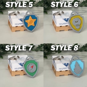 May include: Four different styles of Paw Patrol character badge keychains. Each keychain features a different character's badge with a gray, plastic backing. The badges are yellow, blue, green, and light blue. The keychains are displayed on small wooden crates.
