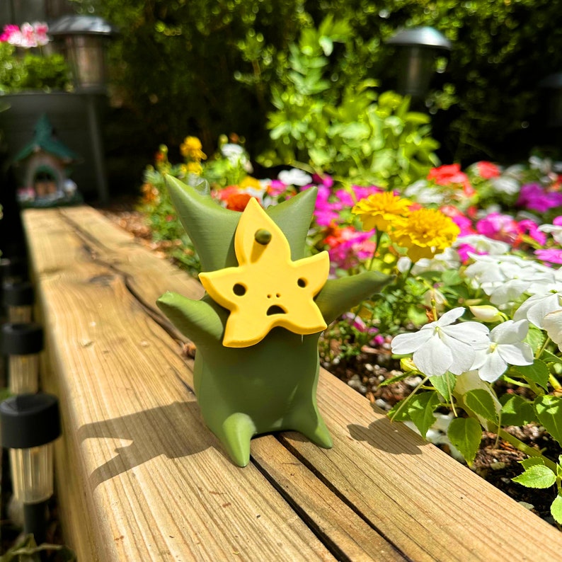 Puede incluir: Una figurita verde con una m&aacute;scara amarilla en forma de estrella, de pie sobre una superficie de madera. La figurita tiene una expresi&oacute;n de sorpresa y est&aacute; rodeada de una variedad de flores.