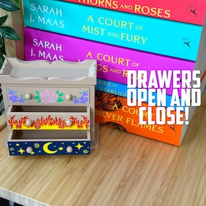May include: A small, beige wooden dresser with three drawers, each decorated with unique designs. The top drawer features a rose and floral pattern, the middle drawer has flames, and the bottom drawer displays a moon and stars. The text "DRAWERS OPEN AND CLOSE!" is visible.