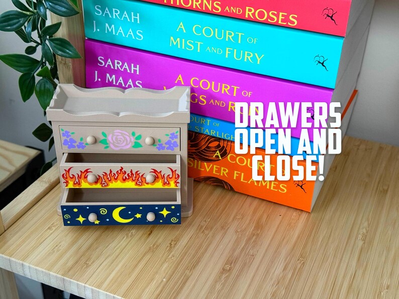 May include: A small, beige wooden dresser with three drawers, each decorated with unique designs. The top drawer features a rose and floral pattern, the middle drawer has flames, and the bottom drawer displays a moon and stars. The text "DRAWERS OPEN AND CLOSE!" is visible.