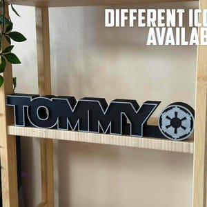 Peut inclure: Une enseigne en bois noir avec le mot "TOMMY" en lettres blanches. L'enseigne est sur une &eacute;tag&egrave;re en bois avec un petit symbole gris de Star Wars &agrave; droite de l'enseigne. Le texte "DIFFERENTS ICONS DISPONIBLES !" est &eacute;crit en lettres blanches sur fond noir au-dessus de l'enseigne.