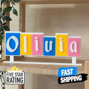 Può includere: Un'insegna con il nome personalizzato "Olivia" in lettere bianche stilizzate. Ogni lettera è su uno sfondo di colore diverso: blu, giallo, rosa. L'insegna è montata su una base bianca, ideale per la cameretta di un bambino.