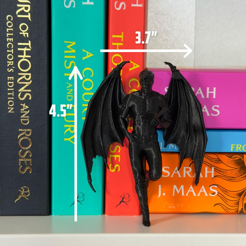 May include: A black figurine of a winged male figure, approximately 4.5 inches tall and 3.7 inches wide, stands in front of a stack of colorful books. The figurine has detailed wings and is posed in a dynamic stance. The books have titles like "A Court of Thorns and Roses."