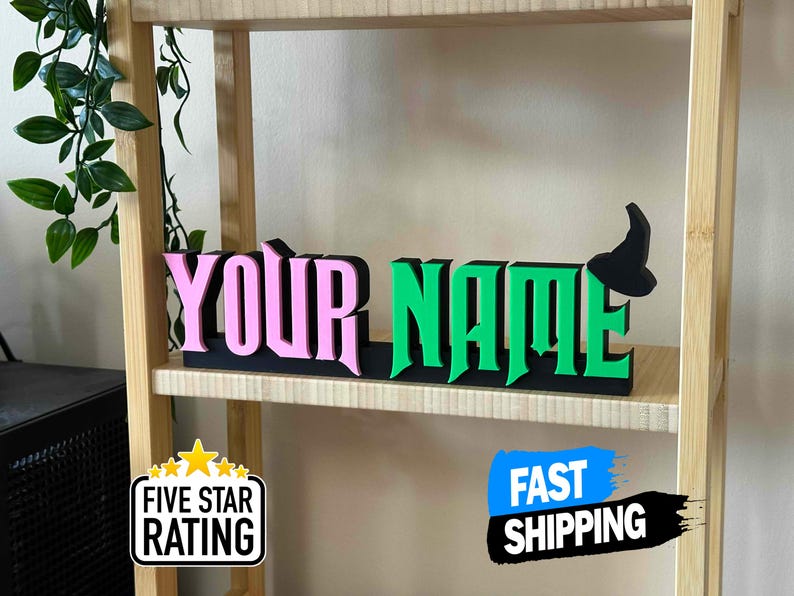 May include: A wooden sign with the words "YOUR NAME" in pink and green letters. The letters are raised and have a 3D effect. A black witch's hat is on the right side of the sign.