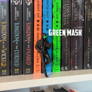 May include: A 3D-printed figurine of a person wearing a green mask and a black outfit, holding a book, stands in front of a row of colorful books. The text "GREEN MASK" is displayed above the figurine.