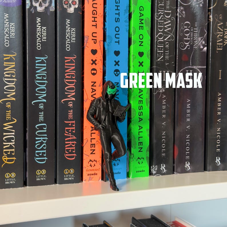 May include: A 3D-printed figurine of a person wearing a green mask and a black outfit, holding a book, stands in front of a row of colorful books. The text "GREEN MASK" is displayed above the figurine.