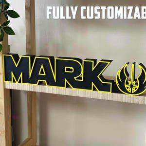 Peut inclure: Une enseigne en bois noire avec une bordure jaune orthographiant "MARK" avec un symbole Star Wars jaune et noir &agrave; droite. L'enseigne est sur une &eacute;tag&egrave;re en bois.