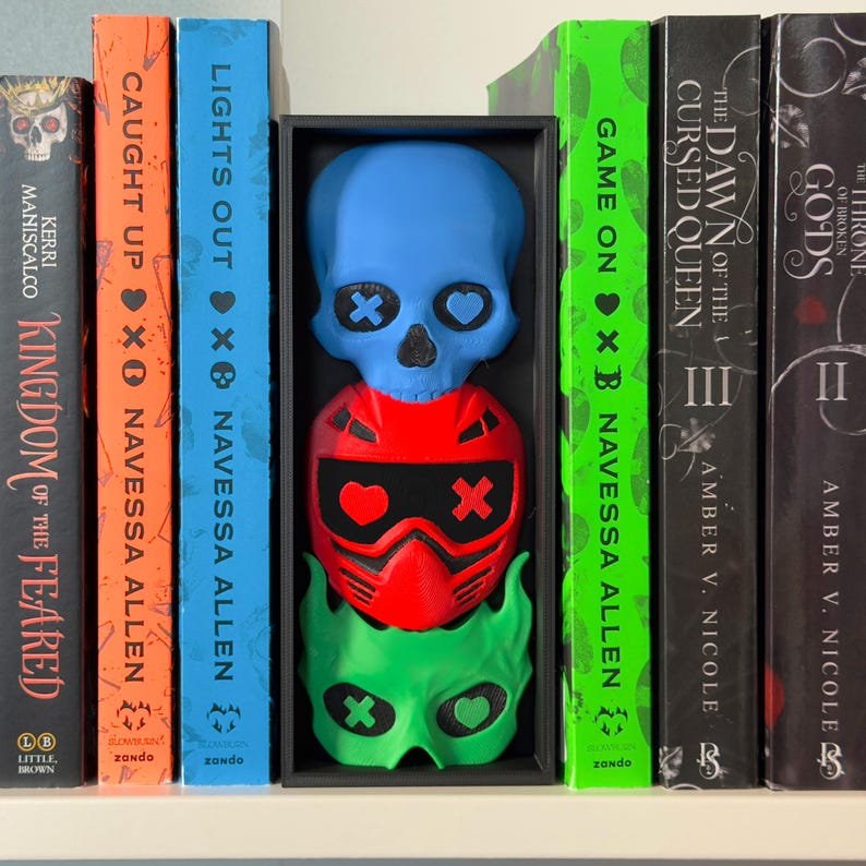 May include: A shelf displays books and a decorative skull sculpture. The sculpture is composed of three stacked, colorful masks: blue skull, red helmet, and green mask. The books have titles like "Kingdom of the Feared" and "Lights Out."