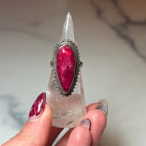 May include: A silver ring with a large, teardrop-shaped, vibrant pink gemstone. The gemstone is set in a detailed, beaded silver frame. The ring is displayed on a clear, pointed stand, showcasing its design and color.