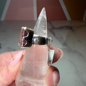 Pu&ograve; includere: Un anello d'argento con una gemma rosa rettangolare. L'anello ha una fascia spessa e un accento nero attorno alla gemma. L'anello &egrave; esposto su un supporto trasparente e appuntito.