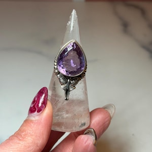 May include: A silver ring featuring a large, pear-cut amethyst gemstone. The amethyst is a vivid purple hue, set in an ornate silver setting. The ring is presented on a clear, cone-shaped display stand.
