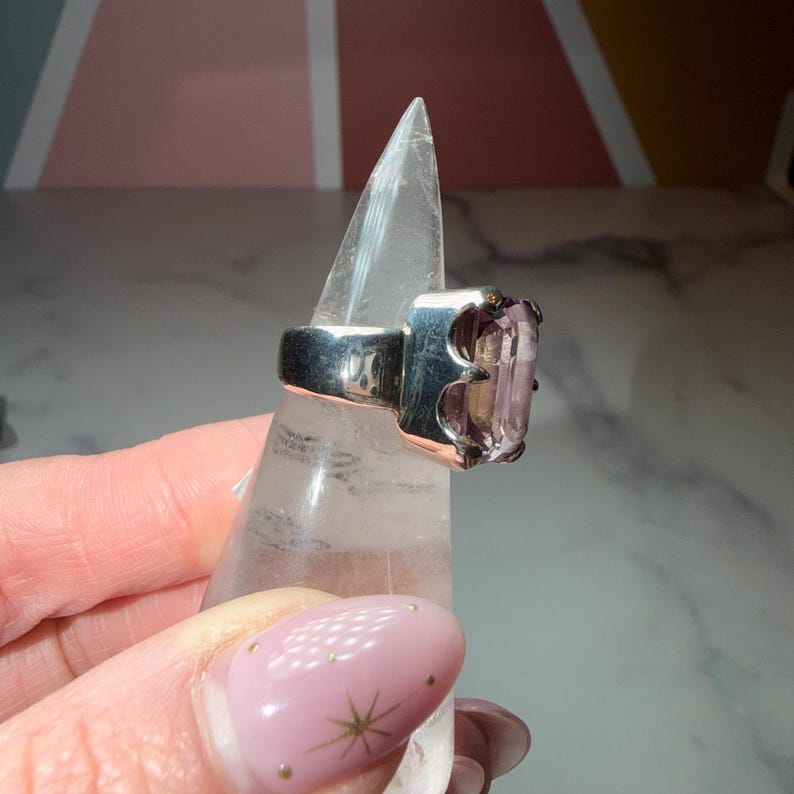 Pu&ograve; includere: Un anello d'argento con una grande gemma rettangolare di colore viola chiaro. L'anello &egrave; esposto su un supporto trasparente e appuntito. La gemma &egrave; il punto focale, evidenziando il suo colore e la sua forma. Il design dell'anello &egrave; moderno e audace.