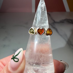 May include: A silver ring with three heart-shaped gemstones in green, orange, and yellow. The ring is displayed on a clear, pointed stand. The ring's design is simple, with the heart-shaped stones set in a silver band.